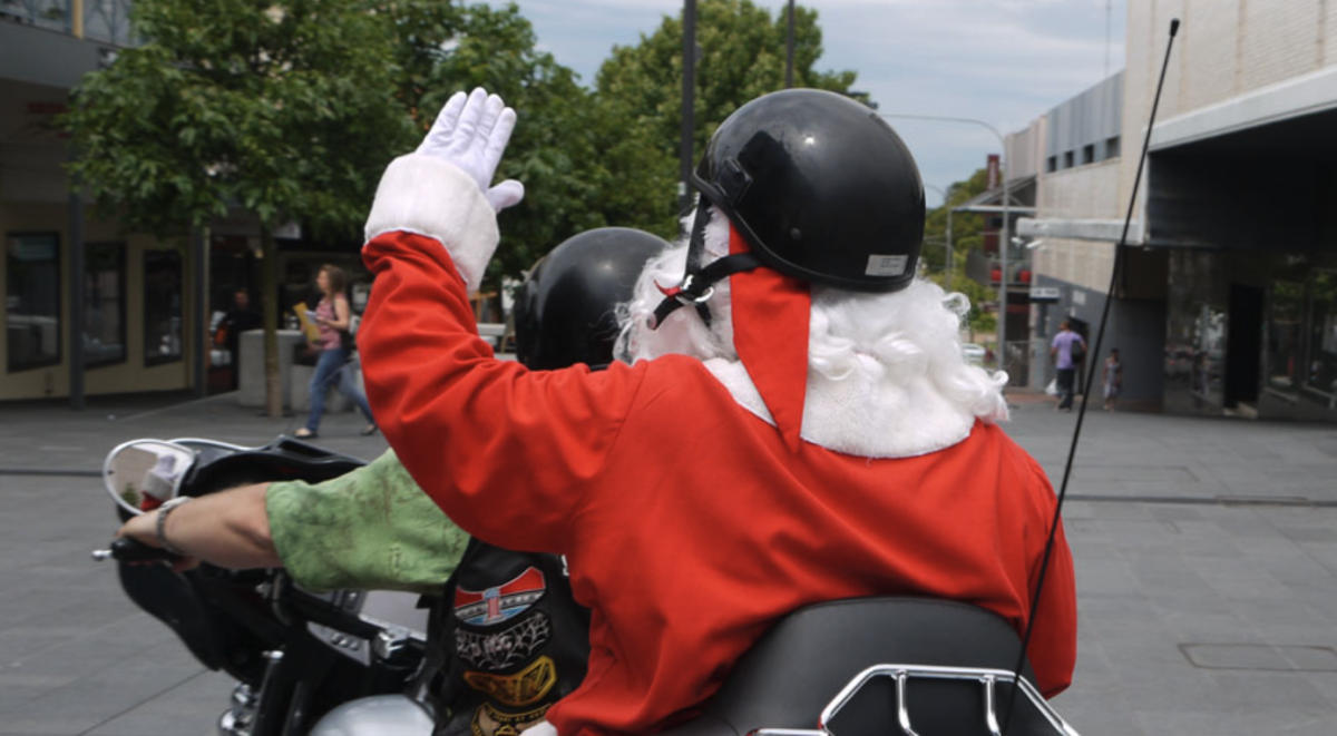 Photo taken from behind of Santa waving as he sits tandem on a motorcycle